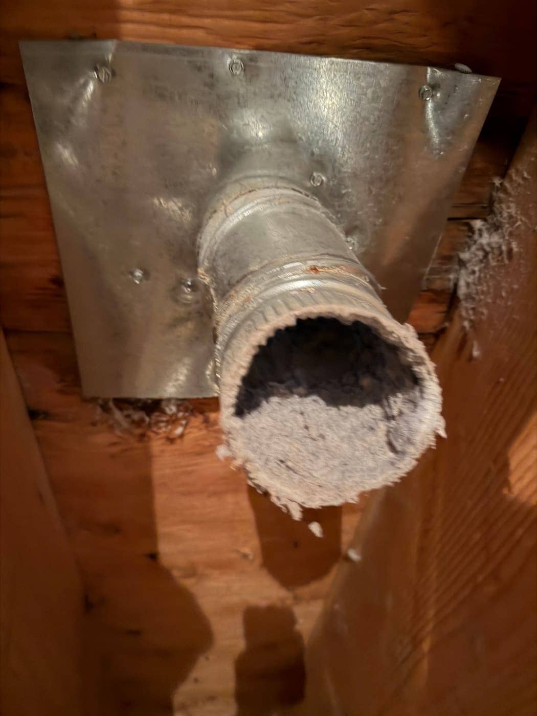 Un conducto de secadora que atraviesa una placa metálica entre vigas de madera, con la boca tapada de pelusa.