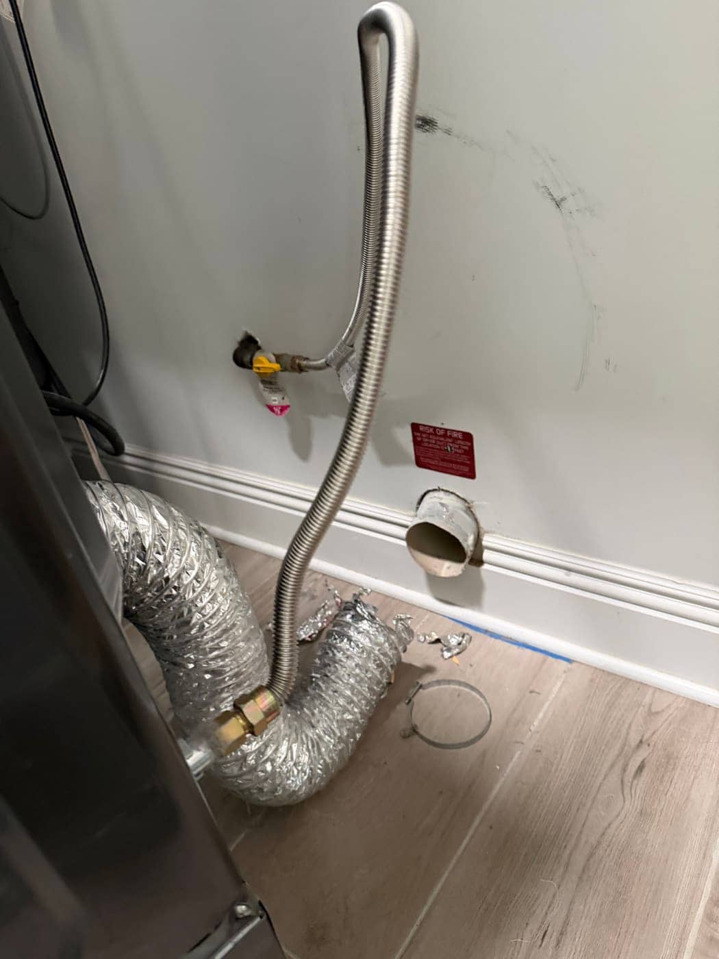 Detrás de una secadora: la línea de gas, un conducto de transición de aluminio arrugado y la salida redonda de ventilación en la pared.